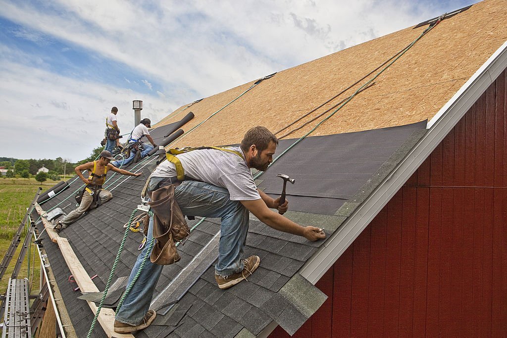A team of men install watreproofing material and shingles to create a new roof on a rural building.